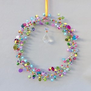 Könnte beinhalten: Ein farbenfroher Perlenkranz mit einer Vielzahl von Perlen in Rosa, Blau, Grün, Gelb und Lila. Ein klarer Kristallprisma hängt in der Mitte. Der Kranz wird von einem gelben Band gehalten.