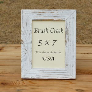 May include: Whitewashed wood picture frame with a distressed finish. The frame is labeled "Brush Creek 5 x 7" and "Proudly made in the USA".