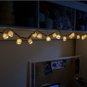 May include: A string of white crocheted lights with a green wire, hanging from a white shelf. The lights are shaped like bells and are lit up, casting a warm glow.