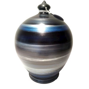Peut inclure: Un pot en céramique avec un couvercle, présentant des bandes horizontales de couleurs dans les tons de bleu, gris et blanc. Le pot a une forme arrondie et un petit bouton sombre sur le dessus. Une petite étiquette est attachée au couvercle.