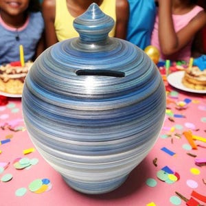 May include: A blue and white striped ceramic piggy bank with a slot for coins. The bank has a rounded shape with a decorative top. The background features a birthday party with balloons and smiling children.