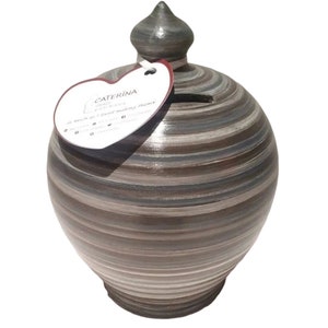 May include: A ceramic piggy bank with a striped pattern in shades of brown, gray, and white. The bank has a rounded body, a small slot for coins, and a decorative top. A heart-shaped tag is attached.