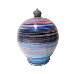 Puede incluir: Una hucha redonda de cerámica con un patrón de rayas en tonos rosa, azul y negro. El banco tiene una pequeña parte superior puntiaguda y una base acampanada. El diseño presenta bandas horizontales de color.