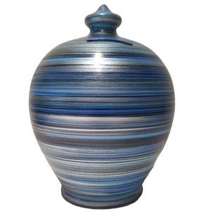 May include: A ceramic money box with a blue and silver striped pattern. The bank has a rounded body, a small, pointed top, and a slot for inserting coins. The colour scheme includes shades of blue, grey, and silver.