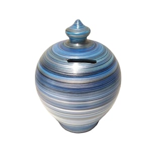 May include: A round, blue and silver ceramic piggy bank with a slot on top for inserting coins. The bank has a decorative, conical lid and is painted with horizontal stripes of varying shades of blue and silver.