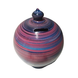May include: A round ceramic piggy bank with a swirl design in shades of pink, blue, and black. The piggy bank has a small opening on the side for coins.