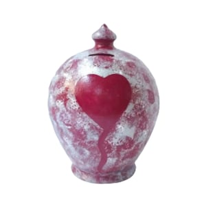 Peut inclure: Une tirelire en céramique avec un motif de cœur rouge. La tirelire est de forme arrondie avec une petite ouverture en haut pour insérer les pièces. Le cœur est peint sur le devant, avec un effet dégoulinant. La tirelire a une finition texturée, mouchetée en rouge et argent.