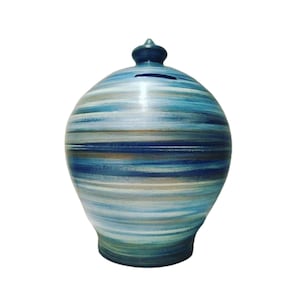 Puede incluir: Una hucha esférica de cerámica con un patrón de pinceladas horizontales en tonos azul marino, azul claro, blanco y marrón tierra. El objeto tiene una pequeña tapa puntiaguda en la parte superior y una ranura horizontal para monedas debajo.