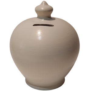 May include: A light beige ceramic piggy bank with a rounded body and a small, decorative top. The bank has a slot for inserting coins and a smooth, glossy finish. The base is a slightly darker shade of beige.