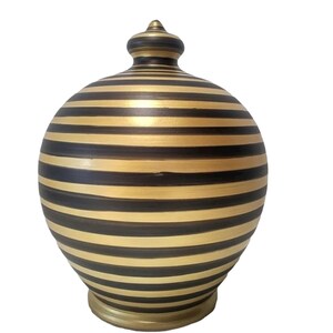 Op de afbeelding: Een decoratieve keramische pot met een deksel, met horizontale zwarte en gouden strepen. De pot heeft een afgeronde body en een kleine, knopachtige finial aan de bovenkant. De basis is ook goud.