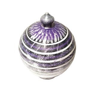 May include: A decorative, spherical jar with a lid. The jar is painted with a pattern of horizontal white stripes, black, and purple. The lid is silver and has a small, rounded finial. The jar is likely ceramic or glass.