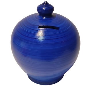 May include: A round, blue ceramic piggy bank with a slot for coins. The bank has a decorative, pointed top and a base. The blue color has a brushstroke effect, creating a textured appearance. The bank is designed for saving money.
