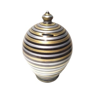 Op de afbeelding: Een bolvormige spaarpot met horizontale strepen in afwisselende metallic goud-, zilver- en donkerbruine kleuren. Het object heeft een puntige knop aan de bovenkant en een smalle horizontale gleuf voor munten in het bovenste gedeelte.