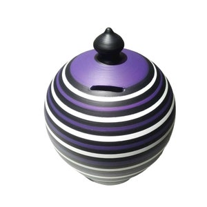 May include: A spherical ceramic bank with a black, purple, and white striped pattern. The bank has a slot for inserting coins and a small, black, conical knob on top. The overall design is modern and decorative.
