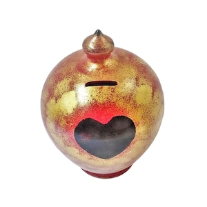 May include: A spherical ceramic piggy bank featuring a metallic gold and red mottled finish. The object has a small pointed top, a horizontal coin slot near the upper section, and a prominent dark heart-shaped cutout on the front surface.