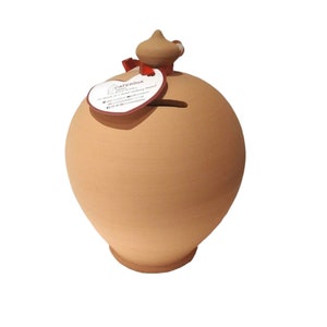 May include: A terracotta piggy bank with a heart-shaped opening and a small lid on top. The piggy bank is decorated with a red ribbon and a white label with the text "CATERINA Handmade Ceramic Pottery".