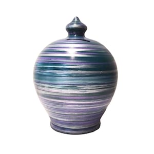 Pode incluir: Um cofrinho redondo de cerâmica com tampa. O banco é pintado com listras horizontais de verde-azulado, roxo e prata. Possui uma fenda para inserir moedas.