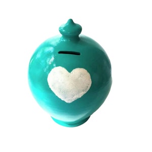 Può includere: Salvadanaio in ceramica color turchese con un cuore bianco dipinto sul davanti. Il salvadanaio ha una fessura nella parte superiore per le monete.