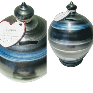 May include: A ceramic money box with a swirl design in shades of blue, grey, and black. The money box has a heart-shaped opening for coins and a silver lid with a pointed top. The money box is decorated with a small white label that says "CATERINA - handmade with love - much as I loved making theme"