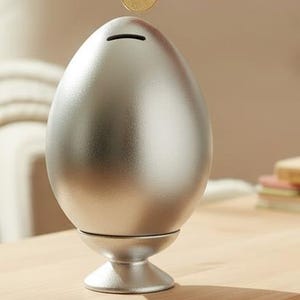 Pode incluir: Um mealheiro em forma de ovo, de cor prateada metálica lisa, com uma ranhura horizontal para moedas no topo, assente numa pequena base circular. O objeto tem um acabamento polido refletor e uma forma arredondada e simétrica.