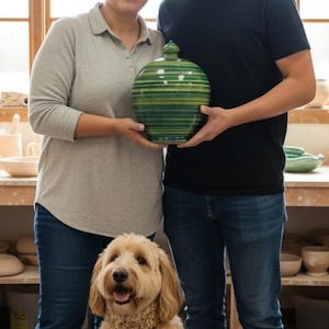 May include: A pottery studio scene featuring two people holding a green and blue striped ceramic vase. A golden doodle dog sits in front of them. Shelves and tables display various ceramic pieces, including bowls, mugs, and vases. Pottery wheels are visible.