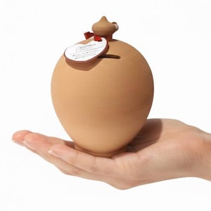 Op de afbeelding: Een kleine, ronde spaarpot van lichtbruin terracotta met een gladde, matte afwerking. Het object heeft een smalle gleuf voor munten aan de bovenkant en een kleine, puntige decoratieve knop. Een wit rond label is bevestigd met een rood lint.