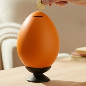 May include: An orange egg-shaped piggy bank with a horizontal coin slot at the top, resting on a black circular pedestal base. The smooth, matte-finished container is designed for saving coins, featuring a simple, rounded form that mimics the shape of an egg.