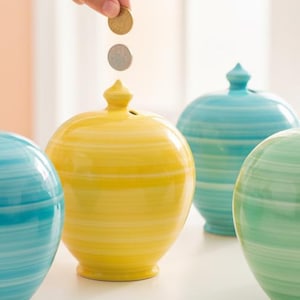 May include: A collection of six colorful ceramic piggy banks in shades of coral, turquoise, yellow, teal, green, and lavender. Each bank has a round shape with horizontal striped detailing and a small, decorative finial on top. Coins are being dropped into the slots.