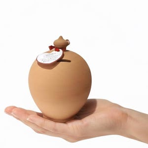 Puede incluir: Una hucha de cerámica marrón claro, con forma de huevo, sostenida en una mano sobre un fondo blanco. El banco tiene un pequeño elemento decorativo en la parte superior y una etiqueta adjunta con una cinta roja.