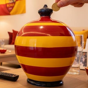 May include: A red and yellow striped ceramic piggy bank with a black top. A person is dropping a coin into the bank. The room has a TV showing a soccer game, a flag, and a scarf, all with the AS Roma team logo.