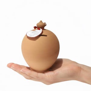 May include: A small, round, light brown terracotta piggy bank with a smooth, matte finish. The object features a narrow coin slot at the top and a small, decorative tag tied with a thin red ribbon around the neck of the opening.