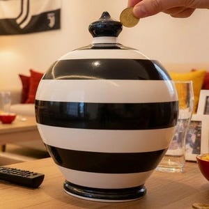 May include: A black and white striped decorative jar with a lid. A person is dropping a coin into the jar. A soccer game is playing on a large TV in the background. A scarf with a soccer team logo is draped over a couch.