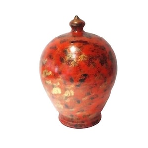 May include: A round ceramic piggy bank with a glossy finish featuring a vibrant red base color, mottled with dark brown and metallic gold patches. The object has a narrow neck, a small pointed lid at the top, and a horizontal coin slot on the front.