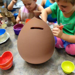 May include: A large, unpainted, brown ceramic piggy bank with a slot for coins. The bank has a rounded shape with a narrow neck and a small opening. Children are painting in the background with colorful bowls and paint.