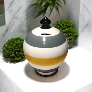 May include: A spherical ceramic piggy bank featuring horizontal stripes in gray, white, and yellow. The bank has a glossy finish, a black pointed lid topper, and a horizontal coin slot located on the upper portion of the rounded body.