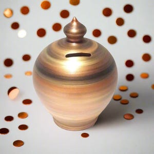 Può includere: Un salvadanaio in ceramica con finitura bronzo metallizzato. La banca ha un corpo rotondo con una fessura per le monete e una parte superiore decorativa. Il colore passa dal bronzo chiaro a quello scuro. Coriandoli color rame sono sparsi intorno alla banca.
