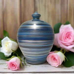 Può includere: Un salvadanaio in ceramica con un motivo a righe blu, grigie e argentate. La banca ha una piccola fessura sulla parte superiore e un terminale decorativo. Rose rosa e bianche circondano la banca.