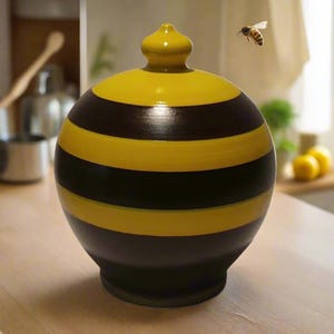May include: A round, hand-painted ceramic jar with a yellow and black striped design, topped with a small, yellow knob. A bee is flying in the background. The jar is approximately 10 inches tall.
