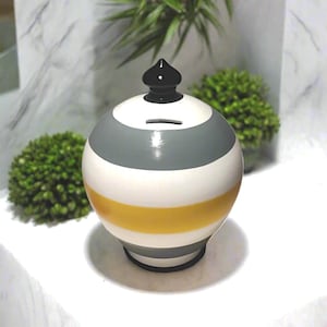 Pode incluir: Um cofrinho de cerâmica redondo com listras horizontais cinza, brancas e amarelas. O cofrinho tem uma base preta e um topo preto e pontiagudo. Uma fenda para inserir dinheiro é visível na parte superior. Um item decorativo.