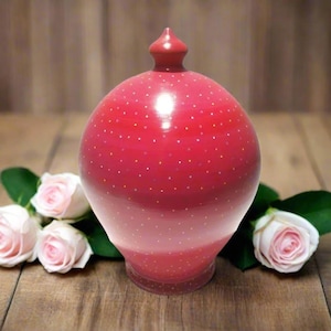May include: A red decorative vase with a small, pointed top and a rounded body, decorated with small white dots. The vase is on a wooden surface, with pink roses and green leaves.