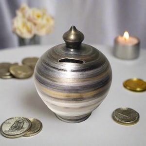 Puede incluir: Una hucha redonda de cerámica con una ranura para monedas. Presenta un acabado metálico con bandas plateadas, doradas y grises. Un pequeño remate a juego corona la hucha. Varias monedas están esparcidas alrededor.