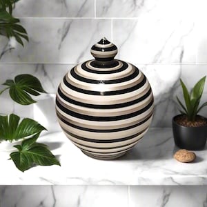 Peut inclure: Un pot décoratif en céramique avec un couvercle, orné de rayures horizontales noires, beiges et blanches. Le pot a une forme arrondie et repose sur une surface blanche. Le couvercle est surmonté d'un petit bouton conique.