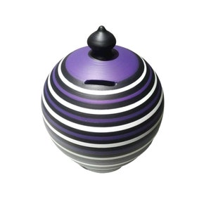 Puede incluir: Una hucha esférica de cerámica con un remate negro y una ranura para monedas. El banco está pintado con rayas alternas de color púrpura, negro, blanco y gris. El diseño es una versión moderna de un artículo clásico.