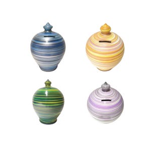 May include: Four ceramic piggy banks with a striped design in blue, yellow, green, and purple. Each bank has a slot for coins and a decorative top. The banks are on a white background.