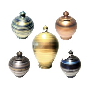 Puede incluir: Colección de cinco huchas de cerámica en varios colores. Las huchas tienen forma redonda con una ranura para monedas y una parte superior decorativa. Los colores incluyen azul, marrón, dorado y plateado. Cada hucha tiene un patrón de rayas único.