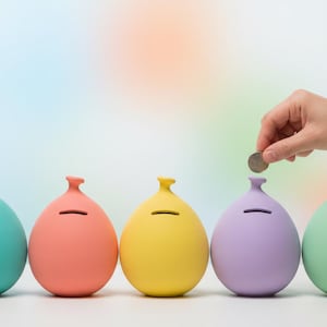 May include: A row of five colorful, egg-shaped piggy banks in teal, coral, yellow, lavender, and mint green. Two hands are shown dropping coins into the slots of the piggy banks. The background is a soft gradient of colors.