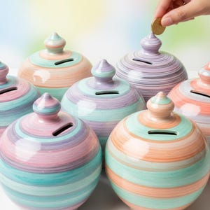 May include: A collection of seven ceramic piggy banks in pastel colors, including pink, blue, and orange, with a striped design. Each bank has a slot for coins and a decorative top. Hands are shown inserting coins into the banks.