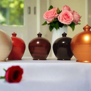 May include: A collection of decorative ceramic vases in various colors, including cream, red, brown, black, and copper. A bouquet of pink roses in a white vase and a single red rose are also present, all arranged on a white surface.