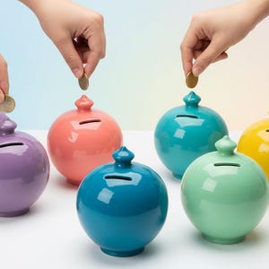 May include: A collection of six spherical ceramic piggy banks in various glossy colors including purple, coral, teal, mint green, and yellow. Each bank features a small top finial and a horizontal coin slot for depositing currency into the container.
