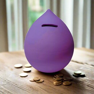 Pode incluir: Um cofrinho de cerâmica roxo em forma de lágrima com uma fenda para moedas no topo. O cofrinho está sobre uma superfície de madeira com várias moedas de ouro espalhadas ao redor.